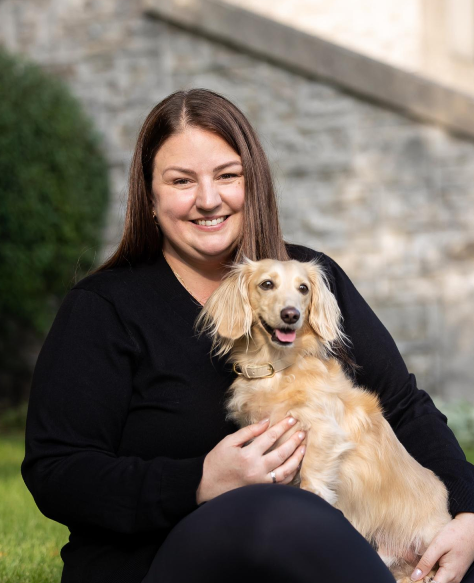 A woman with long brown hair sitting on the grass holding a miniature dachshund on her lap