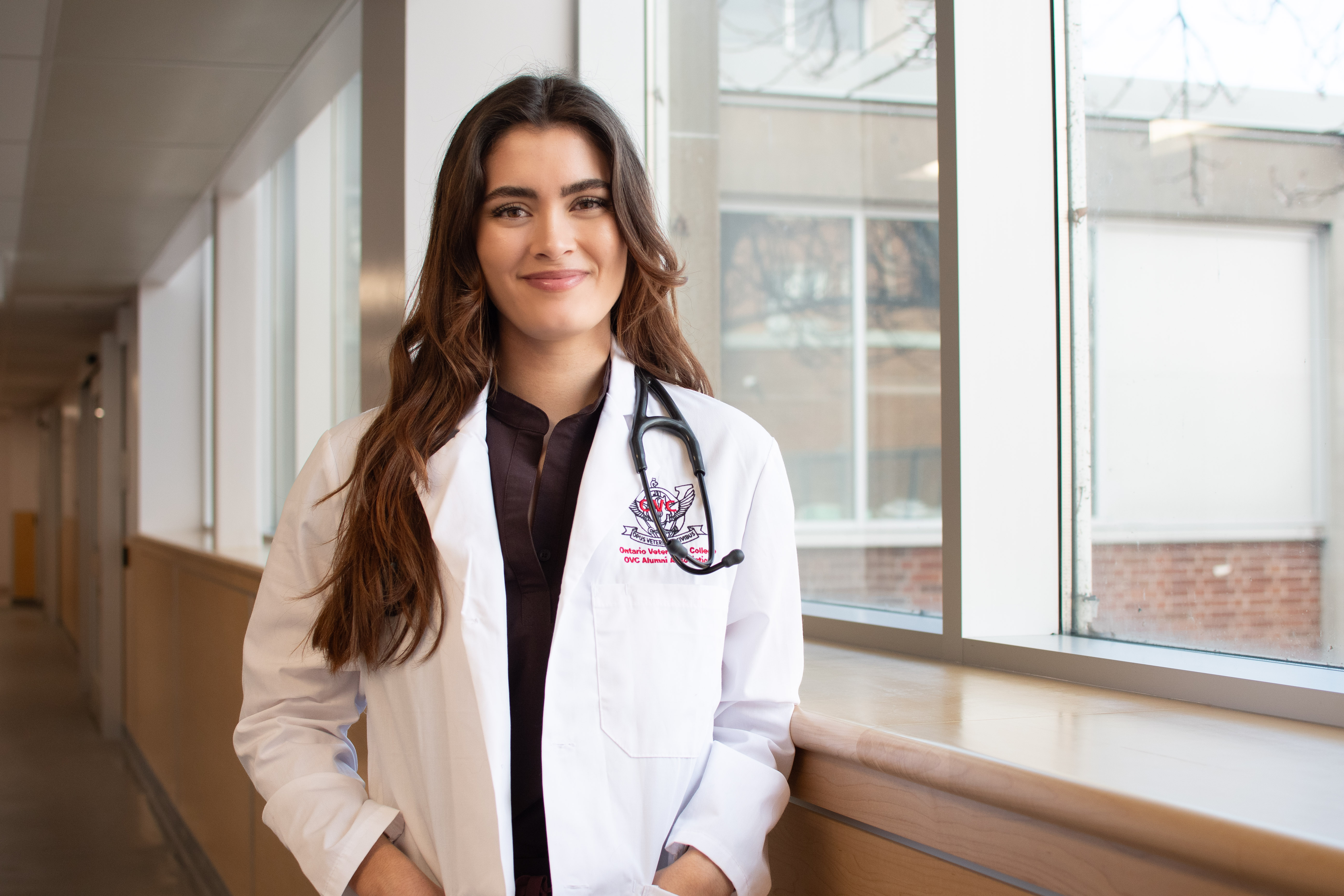 A woman wearing a white Ontario Veterinary College lab coat stands in a bright hallway by a large window. She has long brown hair and a stethoscope draped around her neck.