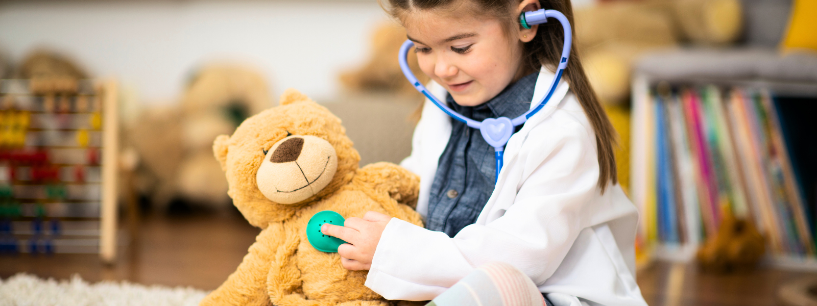 A young girl with dark hair holding a teddy bear and pressing a plastic stethoscope against its chest