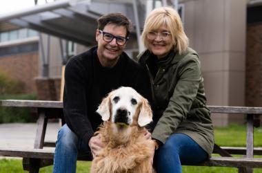 A man and woman sit on a picnic bench with their Golden Retriever dog outside the OVC cancer centre.